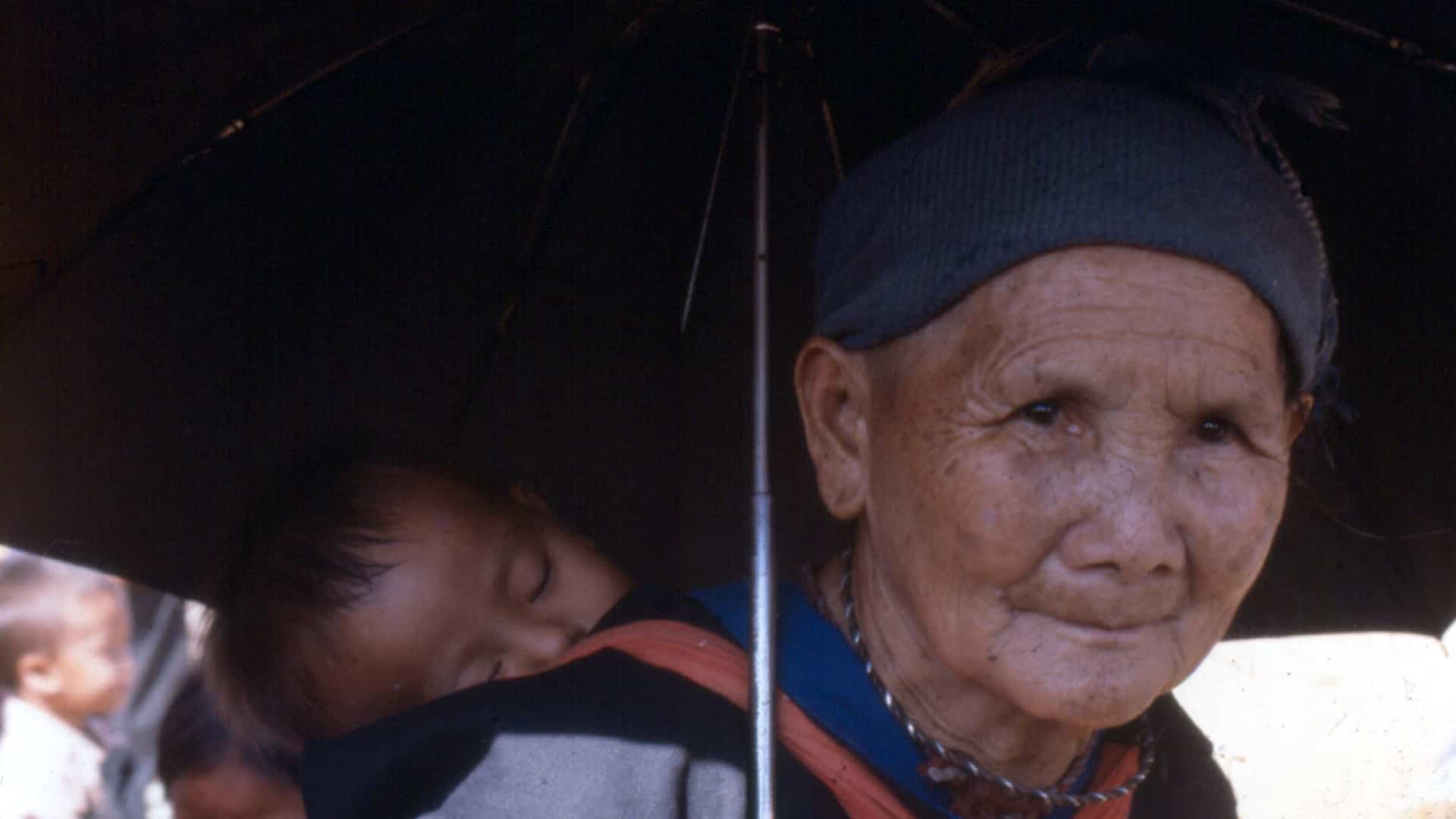 A Hmong grandmother at Ban Vinai camp, a sleeping grandchild carried on her back and two more children beside her